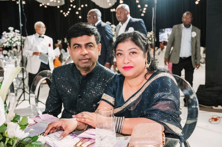 Couple in formal Indian attire posing at an elegant indoor event with decorated tables and guests in the background