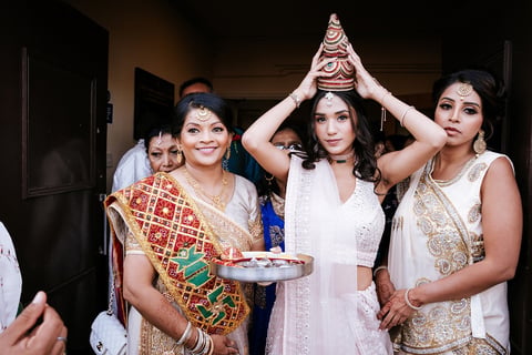 Four women in traditional South Asian attire celebrating together indoors, one wearing a decorative headpiece, holding food and smiling at the camera