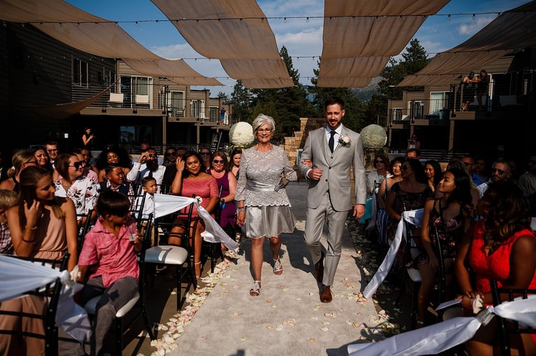 A couple walks down an aisle at an outdoor wedding ceremony with guests seated on both sides under tan shade canopies