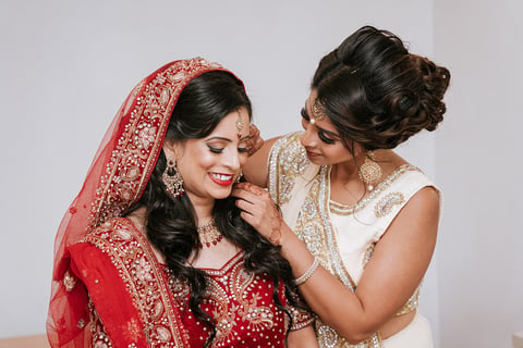 Two women in traditional Indian attire, one wearing a red embellished bridal veil and jewelry, while the other adjusts her veil affectionately