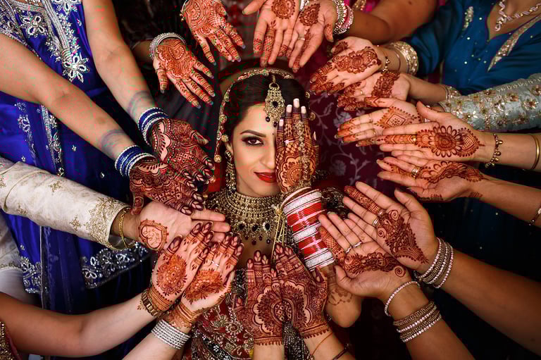 Bride surrounded by female family members displaying henna-decorated hands during Indian wedding celebration