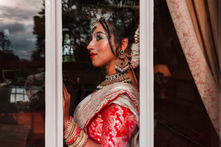 Woman in traditional Indian bridal attire with red garment, jewelry, and makeup gazing out a window