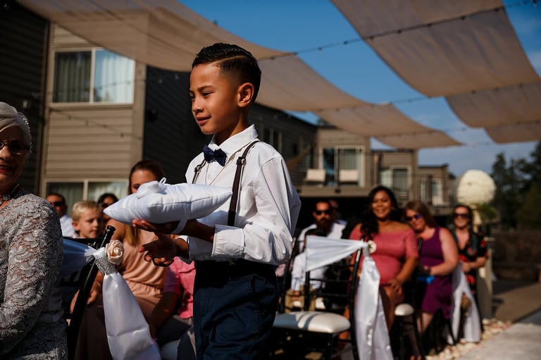 Young boy in formal attire with bow tie gesturing during outdoor celebration with guests and shade structures