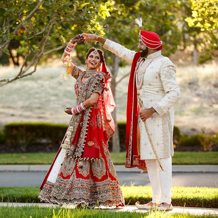 Couple in traditional Indian wedding attire dancing in garden — bride in red lehenga with gold embroidery and groom in white sherwani with red turban — luxury Tanzania wedding photography