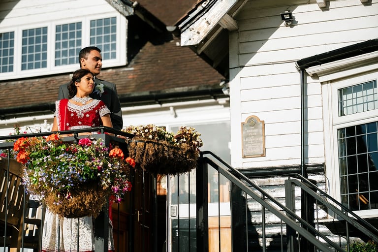 A man in dark clothing and a woman in a red dress stand together on a residential balcony decorated with hanging flowers and plants.