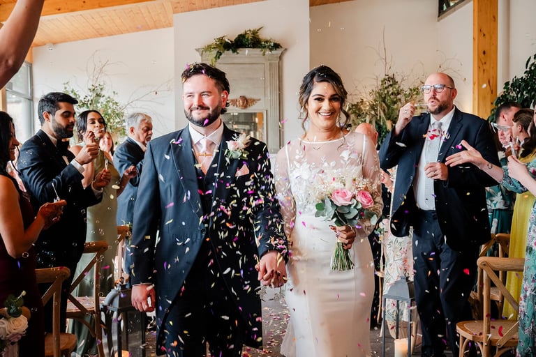 Newlyweds walk down aisle amid confetti, groom in navy jacket with white pattern, bride in white dress holding bouquet, surrounded by cheering guests