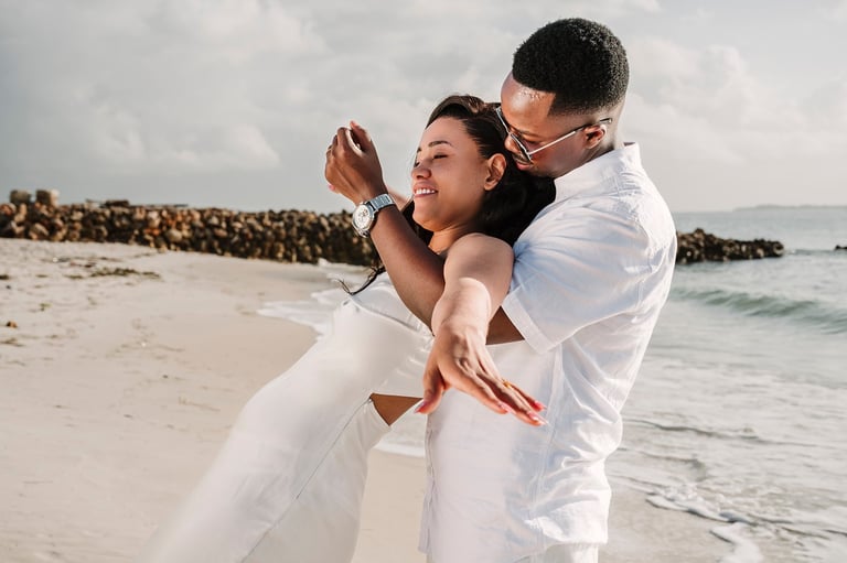 Man carrying woman in white dress on beach, both laughing, with waves and rock jetty in background