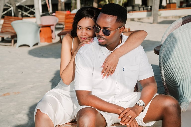 A couple in matching white outfits posing together on a sunny beach with lounge chairs in the background
