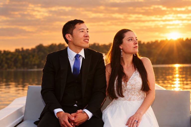 Bride and groom in formal attire sitting on a boat, gazing at a golden sunset over calm water