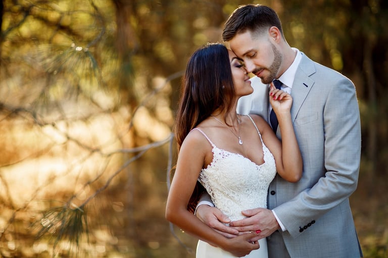 Bride and groom embrace outdoors in golden sunlight, the bride in a white dress and groom in a gray suit among green foliage
