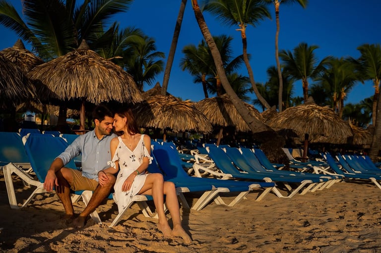 Couple sitting on beach chairs under tiki umbrellas with palm trees and blue sky at sunset
