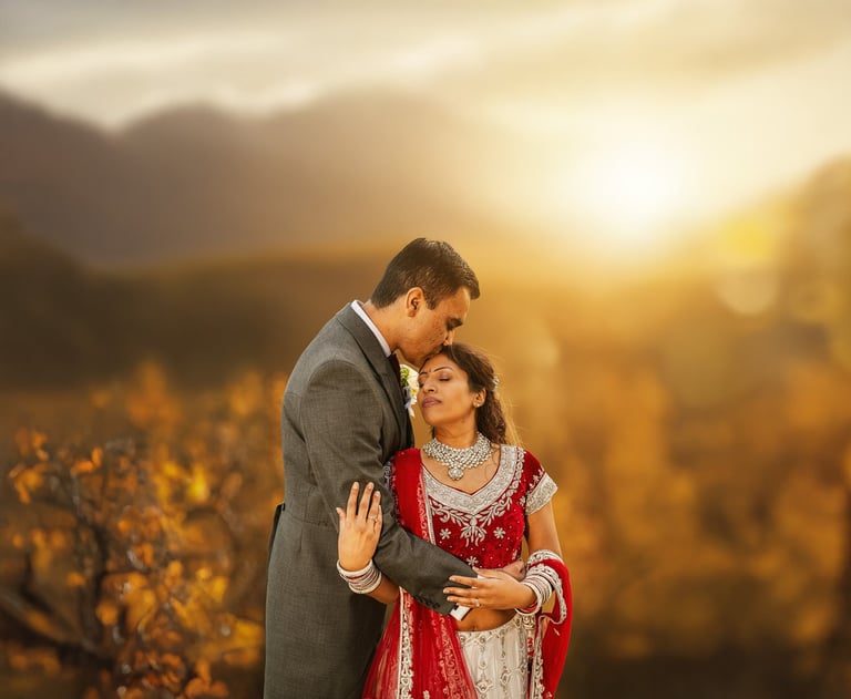 Groom in gray suit kissing brides forehead in red wedding dress in golden sunlit field at sunset