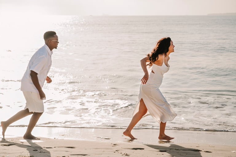 Couple in white clothing running along a sandy beach with ocean waves in the background