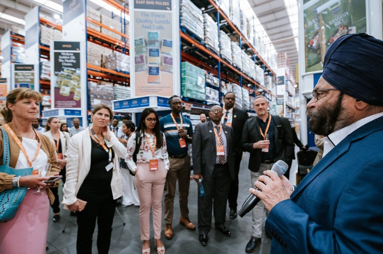 Corporate trade show photography — man in blue blazer addressing group of businesspeople at warehouse trade show with product shelves, Tanzania corporate event