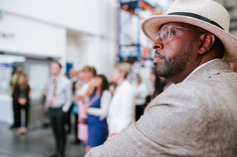 Candid corporate event photography — man wearing straw hat and glasses at indoor business gathering with attendees in background, Tanzania