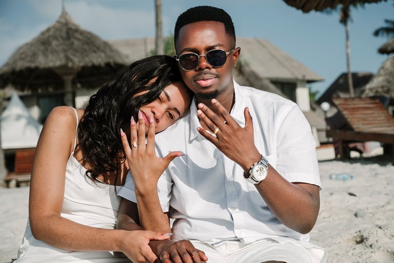 Couple in white clothing posing on a beach with thatched-roof huts in the background