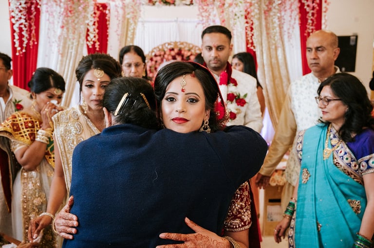 Woman in blue being hugged at Indian wedding celebration with decorated backdrop and guests in traditional attire
