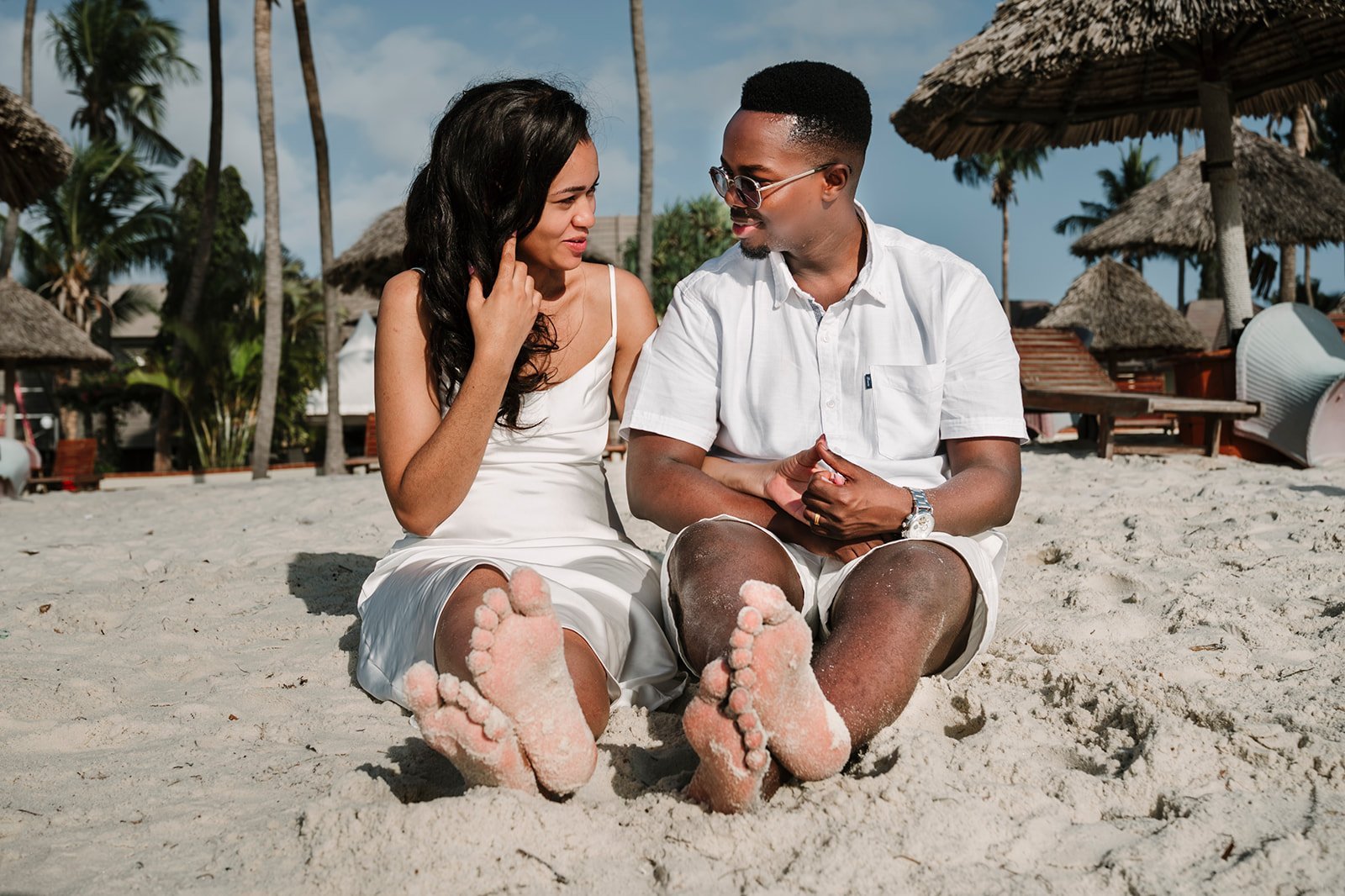 A man and woman sit together on a sandy beach with palm trees and thatched umbrellas in the background